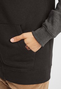Czarna bluza z zamkiem, z szarym rękawem, z kieszenią z przodu i metką. Tkanina wydaje się miękka o gładkiej fakturze.