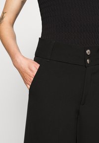 Černé kalhoty s vysokým pasem, které mají hladkou texturu, boční kapsy a kovové knoflíkové detaily v pase.