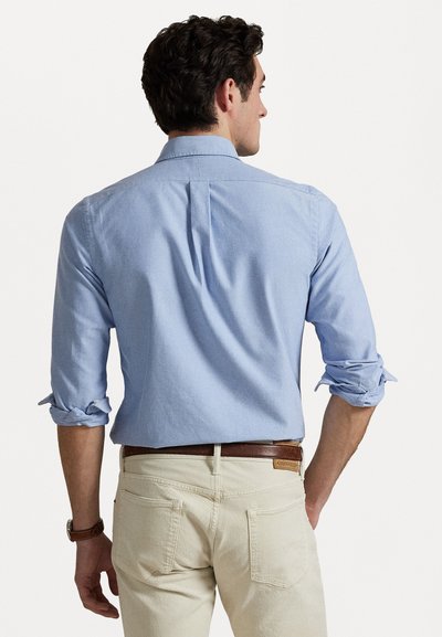 Chemise bleu clair à manches retroussées, col droit et pli au dos, assortie à un pantalon crème et une ceinture marron.
