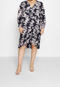 Robe marine à motif floral rose, manches longues, décolleté en V et longueur au genou, associée à des sandales à talons légers roses.