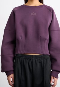 Fialová zkrácená mikina s raglánovými rukávy, s texturovaným logem na hrudi a detailem švů, spárovaná s černými kalhotami na gumu v pase.