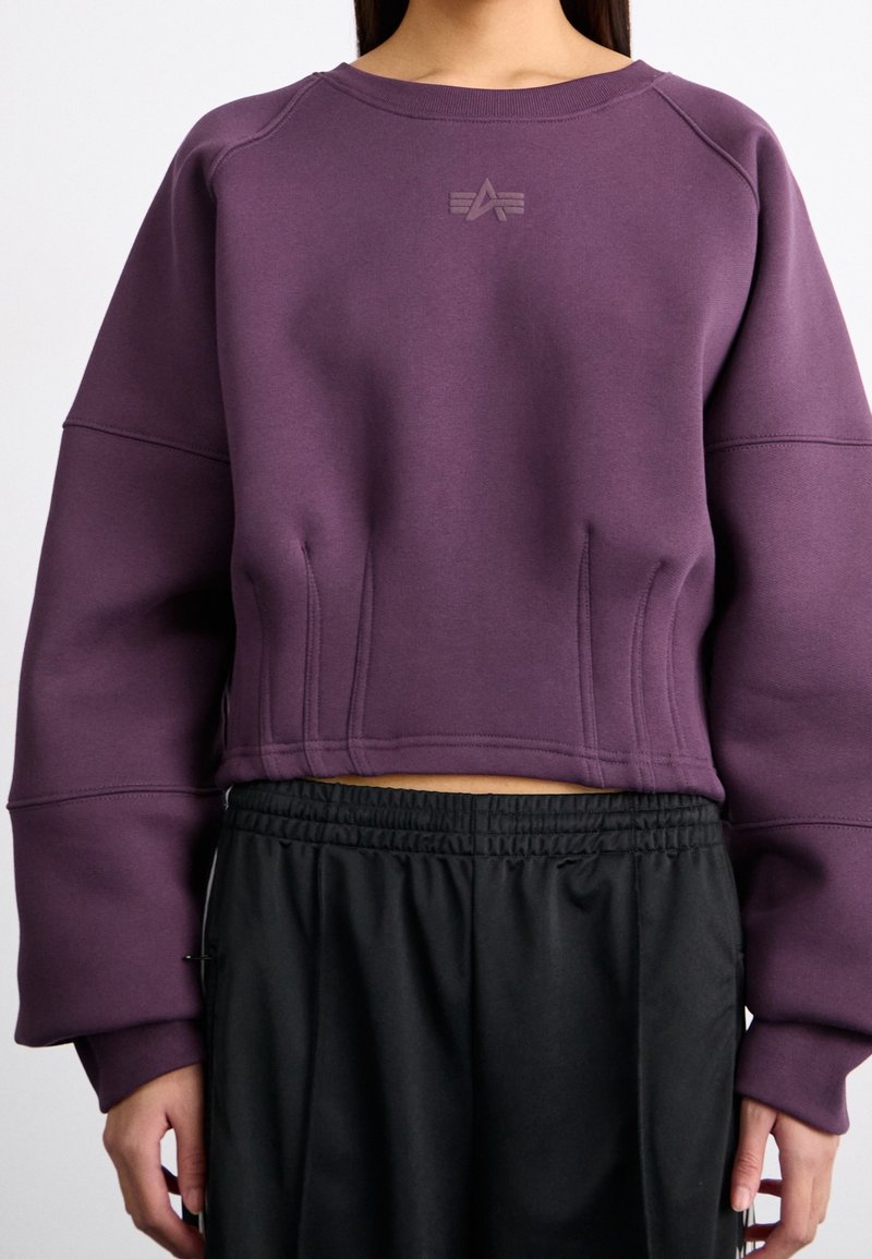 Fialová zkrácená mikina s raglánovými rukávy, s texturovaným logem na hrudi a detailem švů, spárovaná s černými kalhotami na gumu v pase.