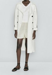 White trench coat with black buttons over a white v-neck shirt, paired with cream knit shorts and black strappy sandals. Smooth textures.