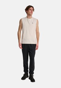Ärmlös krämfärgad bomullsskjorta med en liten svart logotyp på bröstet, kombinerad med svarta joggbyxor och svarta sneakers.