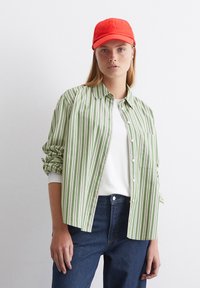 Camicia a righe verdi e bianche con taschino, abbinata a una maglietta bianca semplice e jeans blu. Cappellino rosso con testo ricamato.