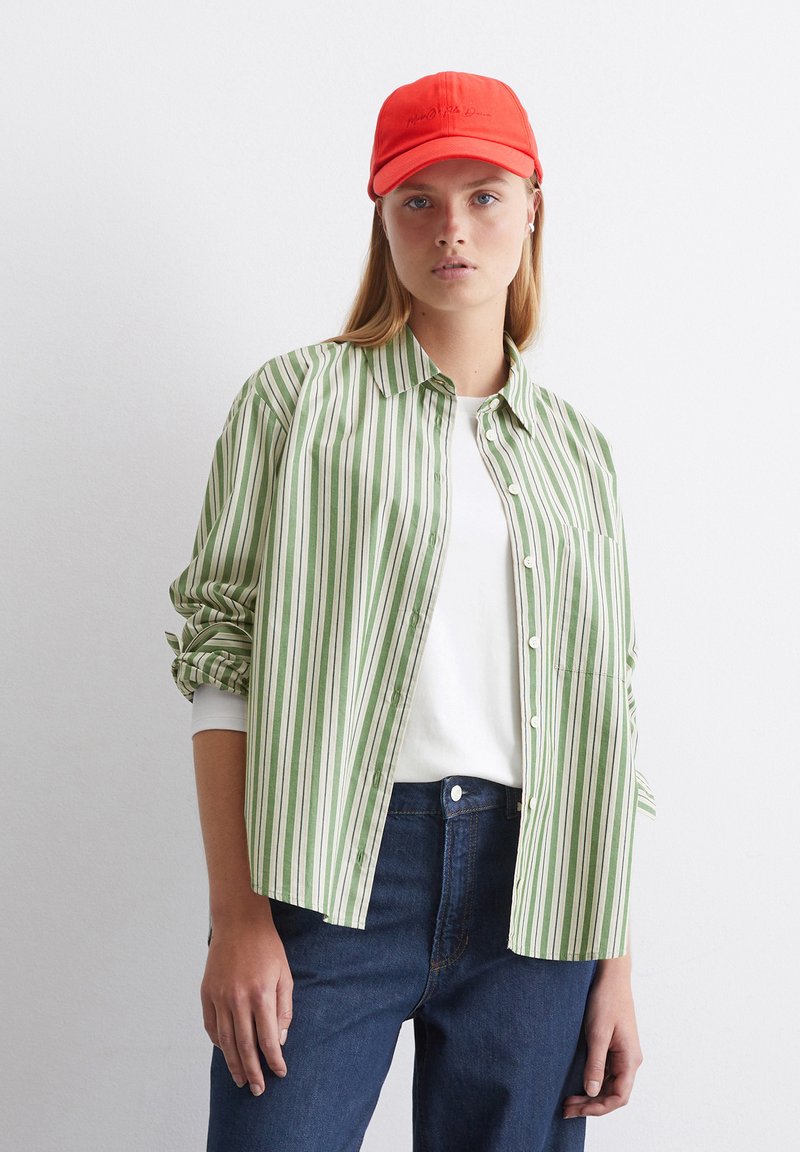Camicia a righe verdi e bianche con taschino, abbinata a una maglietta bianca semplice e jeans blu. Cappellino rosso con testo ricamato.