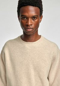 Bēšas krāsas adīta džemperis ar apaļu kakla izgriezumu, fakturētu audumu un brīvu piegriezumu. Materiāls izskatās mīksts un viegls.