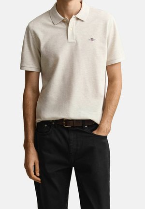 Hombre que lleva una camisa polo de manga corta de color beige claro metida en pantalones negros con un cinturón marrón, una mano en el bolsillo.