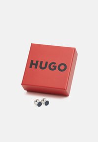 Cureau de argint cu un insert rotund de culoare neagră, prezentat într-o cutie roșie etichetată 'HUGO' cu litere negre îngroșate.