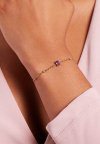 Gouden armband met een paarse edelsteen en meerdere kleine heldere accenten. Fijne kettingontwerp met een gladde textuur. Draagt op een pols.