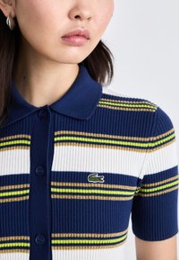 Ribota polo krekls ar svītrām, kas veidots jūras zila, balta un dzeltenzaļa krāsā, ar pogu priekšpusi un izšūtu logo. Īsas piedurknes.