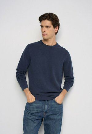 Joven con cabello oscuro, vestido con un suéter azul marino y vaqueros azules, está de pie con las manos en los bolsillos, mirando ligeramente hacia un lado contra un fondo liso.