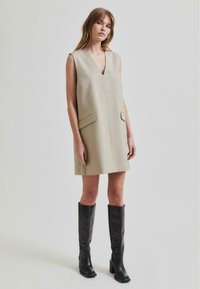 Beige ermeløs kjole med dyp V-hals, to frontlommer og rett snitt, kombinert med høye svarte skinnstøvler.