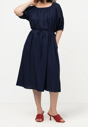 Woman wearing a navy blue off-shoulder midi dress with a waist tie and red woven high-heeled mules, standing against a plain white background.