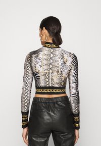 Long-sleeve cropped top with mixed animal prints in beige, black, and gold. Features a high neckline and gold chain accent at the hem.