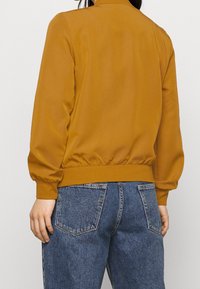 Musztardowa kurtka bomberka z żebrowanym kołnierzem i mankietami, elastycznym pasem oraz balonowymi rękawami, zestawiona z niebieskimi dżinsami.