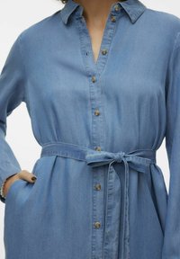 Abito camicia in denim azzurro chiaro con collo a revers, chiusura con bottoni, vita con cintura e tasche laterali. Texture liscia e design casual.