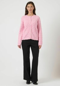 Lys pink strikket cardigan med et struktureret mønster, scallop-kant og rund halsudskæring, med hvide knapper foran. Sorte bukser.