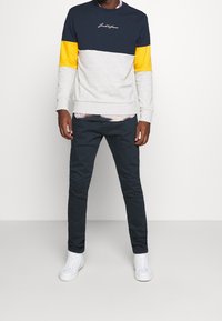 Hombre con sudadera a bloques de color en secciones de azul marino, gris y amarillo, pantalones azul marino y zapatillas blancas, de pie frente a un fondo blanco.