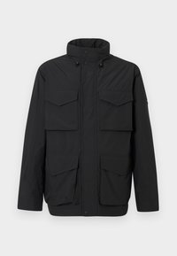 Musta eristetty takki korkealla kauluksella, etuvetoketjulla, nepparilla ja neljällä läpällisellä taskulla vaalealla taustalla.