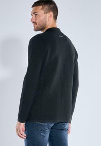 Mann, der über die Schulter schaut, trägt einen strukturierten schwarzen Pullover und eine blaue Jeans und steht vor einem einfarbigen, hellgrauen Hintergrund.