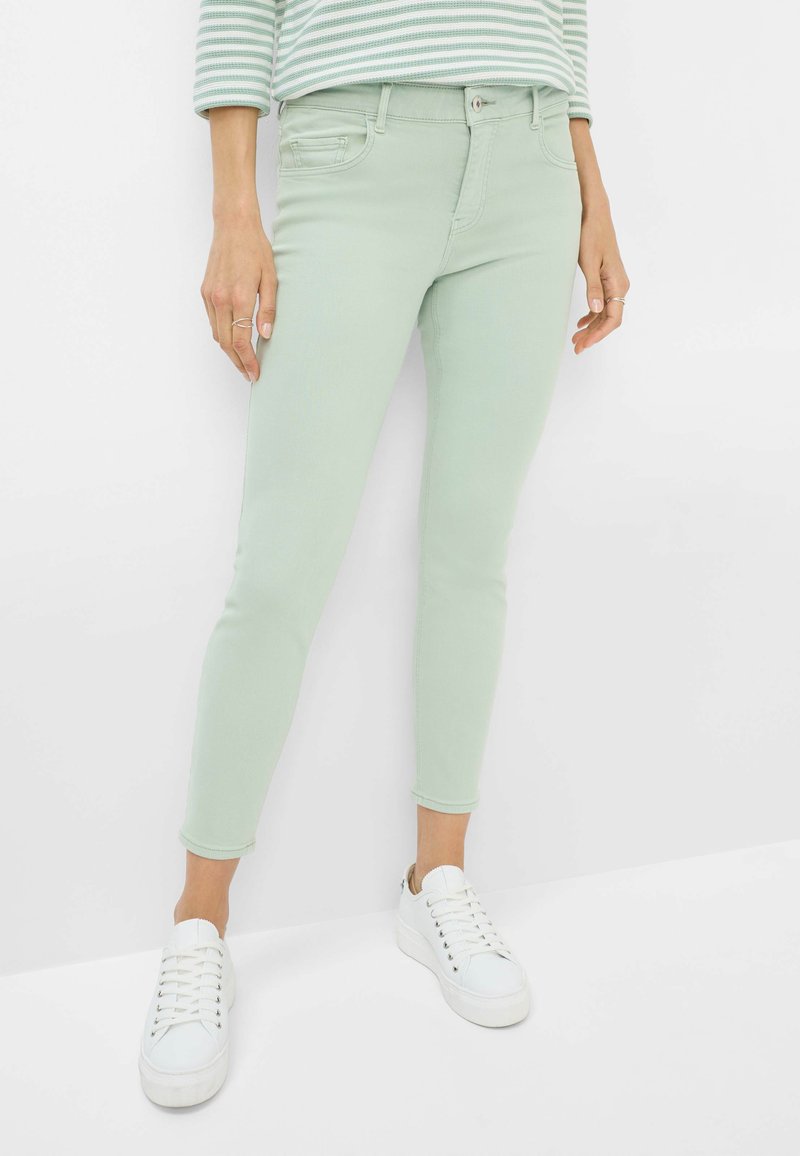 Jeans skinny verde menta con una texture liscia, dotati di una tasca frontale, passanti per cintura e chiusura a bottone. Abbinati a sneakers bianche.