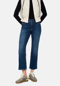 Leichte gesteppte Weste mit Wellenmuster, schwarzes Langarmshirt, dunkelblaue verkürzte Jeans und Sneakers mit Zebramuster und beigen Akzenten.