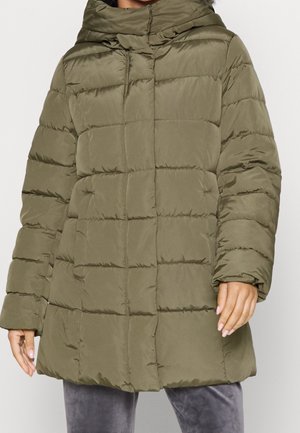 Femme portant une doudoune matelassée vert olive avec un col montant et des poches avant, associée à un pantalon en velours gris.