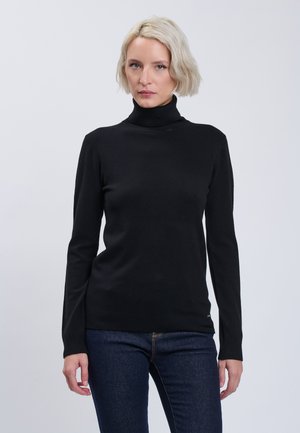Femme blonde aux cheveux courts portant un pull à col roulé noir et un jean bleu foncé, debout devant un fond clair uni.