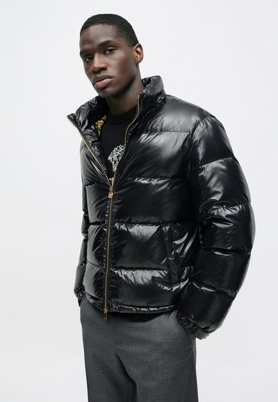 Sort dunjakke med skinnende finish, med en guldfarvet lynlås og høj krave, stylet over en sort grafisk sweater og grå bukser.