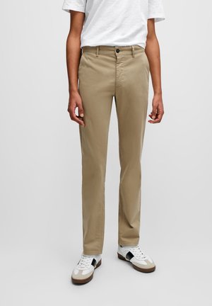 Beige chino pants made of cotton with a slim fit. Features a button closure and pockets, paired with white sneakers with black accents.