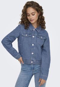 Veste en denim bleu coupée avec poches avant, boutons en argent et col classique. Texture lisse avec une coupe décontractée.