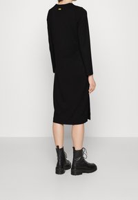 Černé šaty s dlouhým rukávem a uvolněným střihem, které mají boční rozparky a malou žlutou visačku vzadu. Doplněné černými šněrovacími kotníkovými botami.