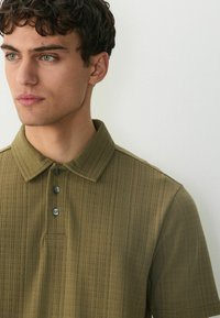 Olivengrøn polo t-shirt med et struktureret mønster, der har en folded krave og en tre-knaps lukning. Korte ærmer og en afslappet pasform.