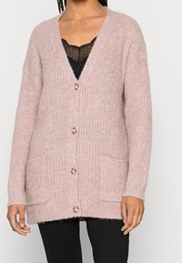 Žena v růžovém silném pleteném cardigan s knoflíky vpředu a kapsami na nášivky, oblečená přes černý top s krajkovým lemem a černé kalhoty.