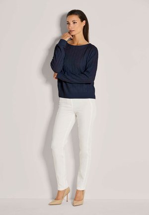 Eine Frau steht an einer Wand, trägt einen marineblauen, grob gestrickten Pullover, weiße enge Hosen und beige High Heels, mit der Hand nahe am Gesicht.