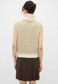 Gilet beige lavorato a maglia senza maniche con colletto alto a coste e fondo a coste, abbinato a una gonna marrone plissettata.