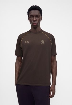 Jeune homme aux cheveux courts et bouclés porte un t-shirt marron EA7 avec logos et un pantalon foncé sur un fond blanc uni.