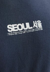 Marine T-shirt med fremtrædende hvid tekst, der står "SEOUL", samt stedsnavne på flere sprog, på et struktureret bomuldsstof.