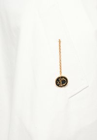 Zelta ķēde ar melnu un zeltu ovālu piekariņu, kas karājas no baltas auduma ar šūtu vāku.