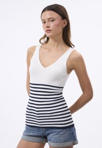 Top a canotta a coste con scollo a V profondo in bianco, caratterizzato da righe orizzontali blu navy e bianche sulla metà inferiore, abbinato a pantaloncini di jeans.
