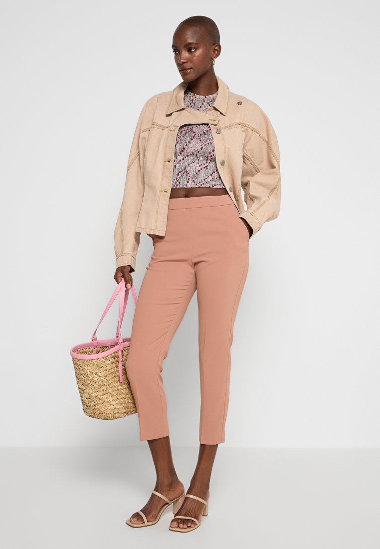 Beżowa dżinsowa kurtka, krótki wzorzysty top, różowe dopasowane spodnie i beżowe sandały. Trzyma plecioną torbę ze słomy z różowymi akcentami uchwytów.