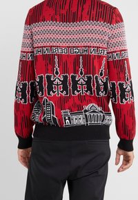 Roter gestrickter Pullover mit schwarzen und weißen Stadtansichten und Musterdesigns, mit gerippten Bündchen und Saum. Das Textil hat eine traditionelle Ästhetik.