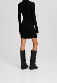 Robe noire côtelée à manches longues avec une silhouette ajustée, longueur au-dessus du genou, associée à des bottes noires brillantes montant jusqu'au genou.