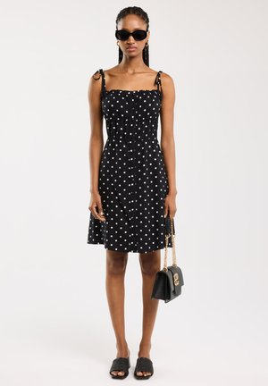 Schwarzes Kleid mit Punkten, Spaghetti-Trägern, gesmoktem Oberteil und weit schwingendem Rock. Dazu passende Accessoires sind eine schwarze Handtasche und Sonnenbrille.