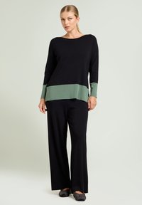 Zwarte gebreide top met een groene onderrand en mouwen, gecombineerd met zwarte wijde broek. Glad textuur, relaxte pasvorm en een eenduidig kleurenpalet.