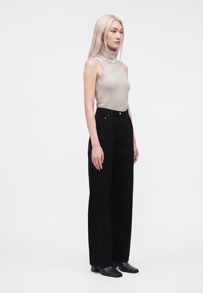 Jeune femme aux longs cheveux blonds portant un haut rayé sans manches à col montant, un pantalon noir à jambes larges et des bottines noires, debout de profil.
