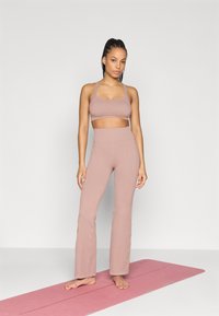 Σετ αθλητικών ρούχων σε ανοιχτό ροζ χρώμα, που περιλαμβάνει εφαρμοστό αθλητικό σουτιέν και ψηλόμεσα κολάν με φουσκωτή γραμμή, κατασκευασμένα από λείο ελαστικό ύφασμα, πάνω σε στρώμα γιόγκα.