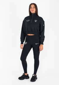 Felpa nera corta con zip a metà; presenta il logo dei Las Vegas Raiders. Leggings neri con pannelli laterali in rete e logo Nike. Scarpe da ginnastica nere.