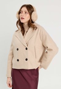 Manteau en laine beige à double boutonnage avec de grands boutons noirs, poches latérales et un design avec col. Associé à un pull à col haut et une jupe bordeaux.